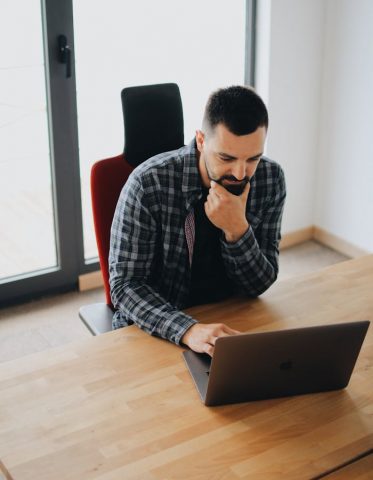 Young man with beard working on laptop at desk in a modern office setting.